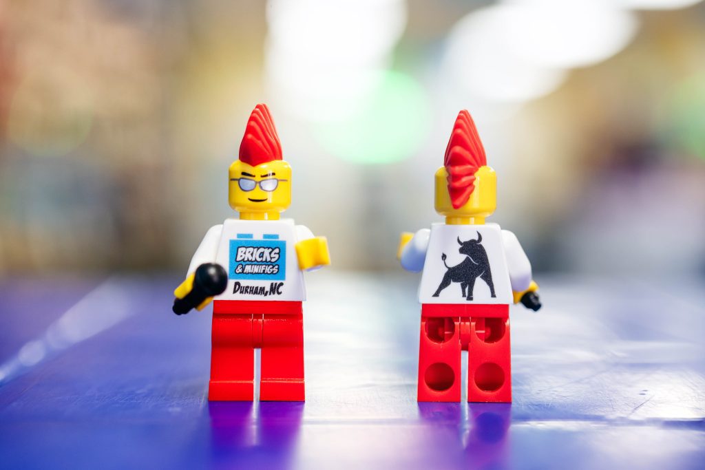 A LEGO minifigure with a bright mohawk, featuring the Bricks & Minifigs logo on its front and a Durham bull graphic on its back.