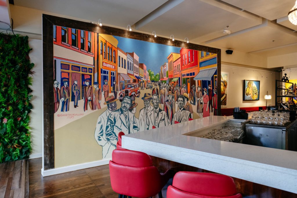 A colorful mural depicting Durham’s historic Black Wall Street stretches behind the bar at Flavor Hills Restaurant & Bar, with red barstools and a white countertop in the foreground.