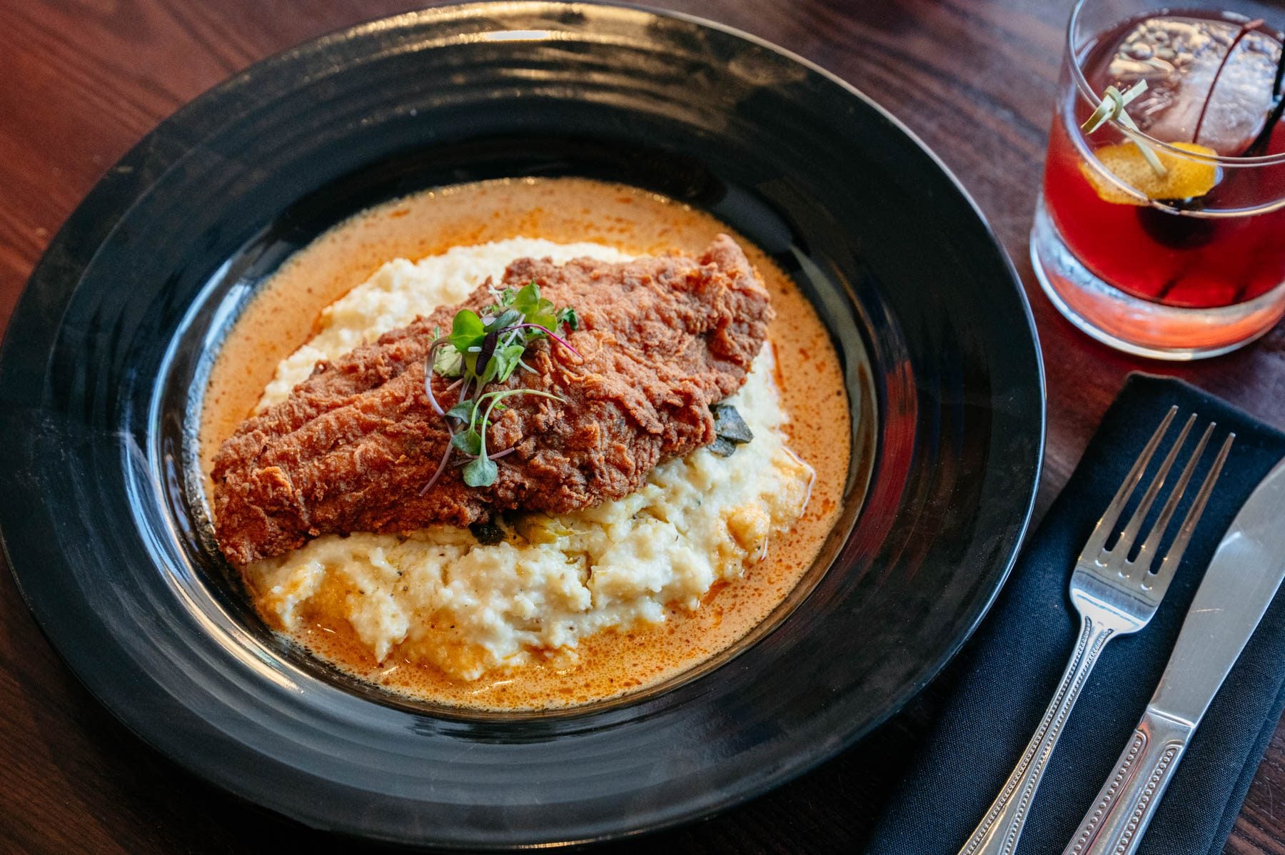 A piece of golden fried catfish sits atop creamy cheese grits with collards and Cajun cream sauce, served in a black bowl beside a cocktail.