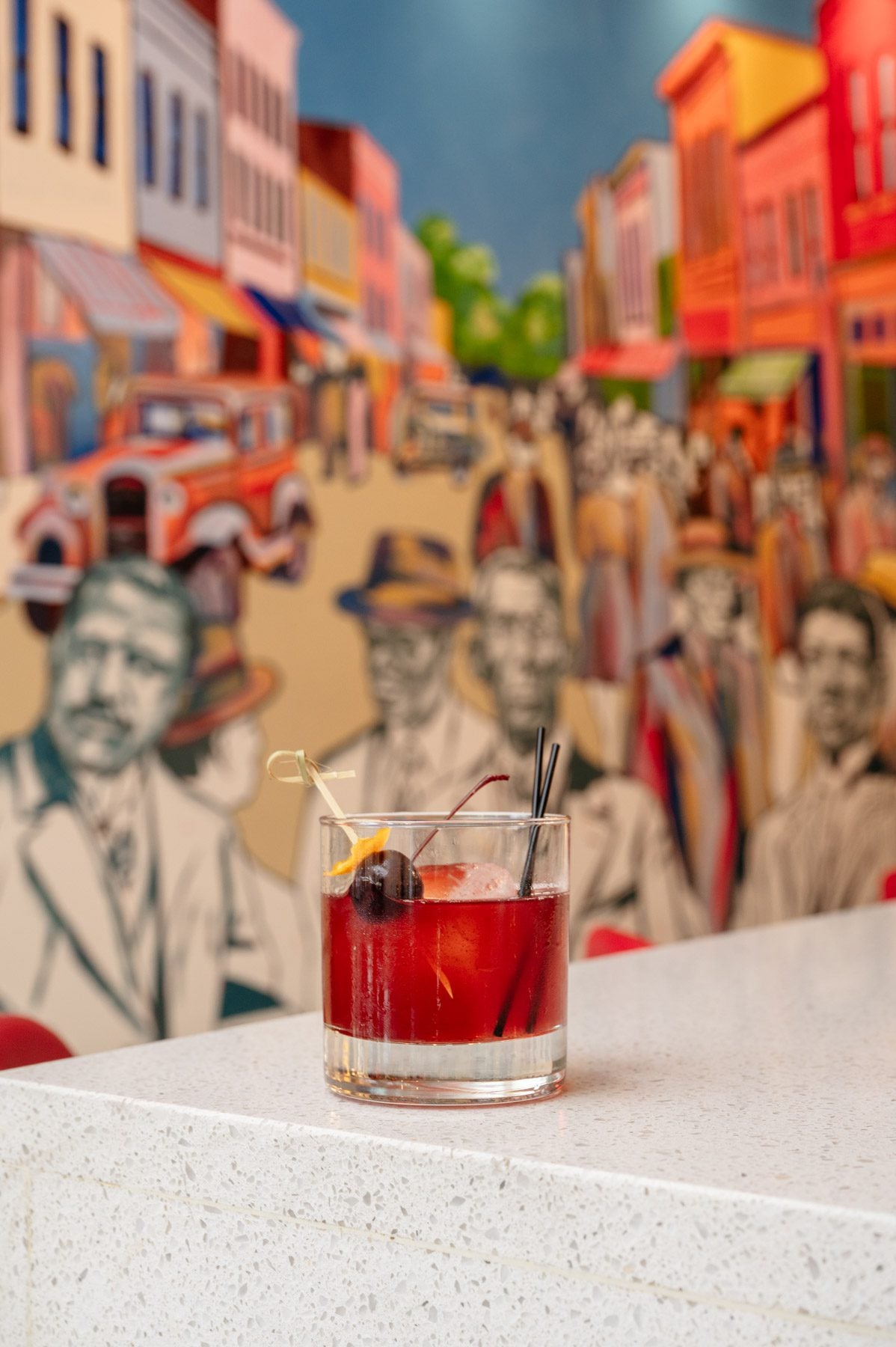 A blackberry old-fashioned cocktail garnished with a cherry and orange peel sits on a bar counter in front of a colorful mural depicting Durham’s historic Black Wall Street.