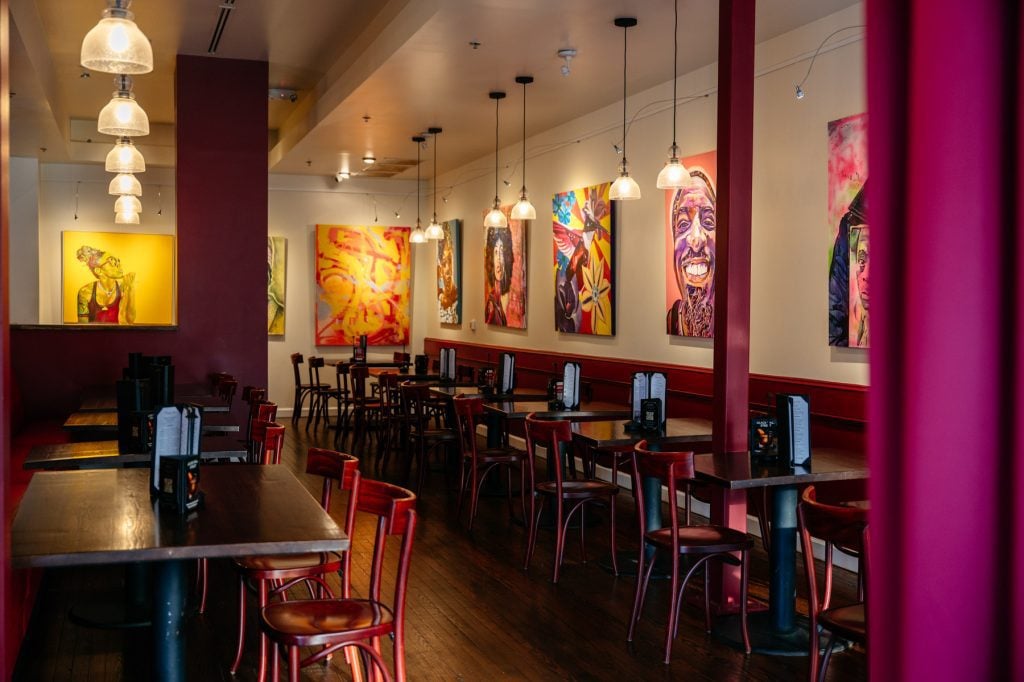 The dining room of Flavor Hills Restaurant & Bar features tables and chairs beneath warm pendant lighting, with vibrant portrait paintings displayed along the walls.