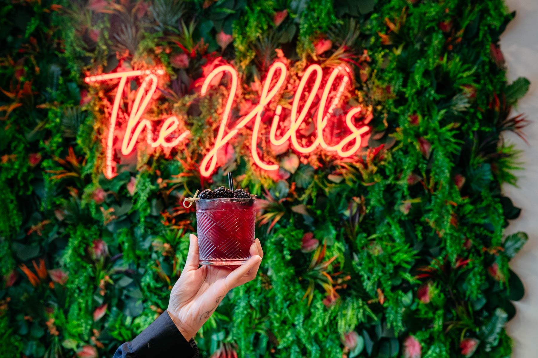 A hand holds a blackberry cocktail in front of a green plant wall with a red neon “The Hills” sign at Flavor Hills Restaurant & Bar.