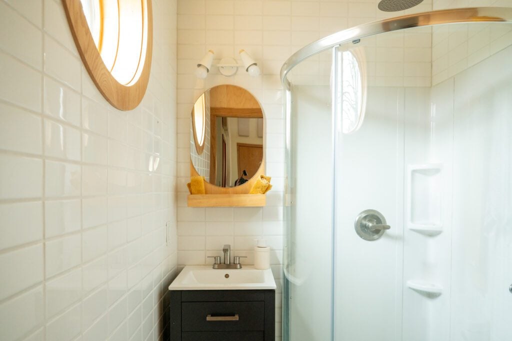 Compact bathroom with a shower, white tile walls, a mirror and a round window resembling a porthole.
