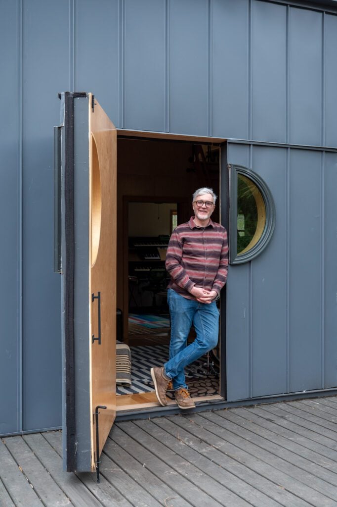 James Phillips standing in the doorway of his modern backyard recording studio with a wooden navy door and a round window.