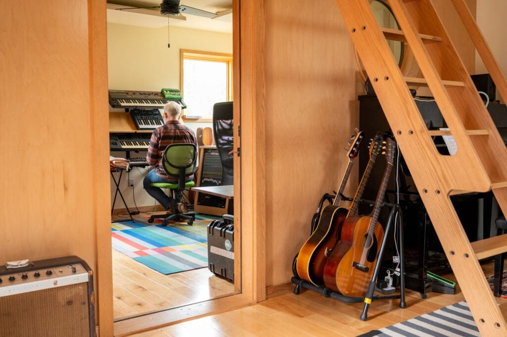 View into a recording studio with a musician at a keyboard, guitars nearby and a wooden ladder leading to a loft.