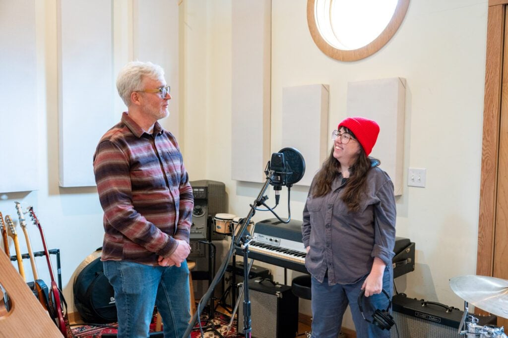Christy Smith stands at a microphone in a recording studio, with James, preparing to record vocals.