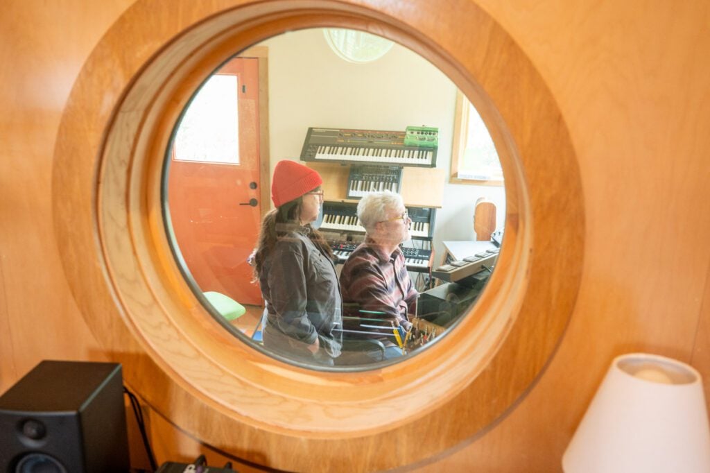 View through a round interior window into a recording studio control room with two people at a desk.