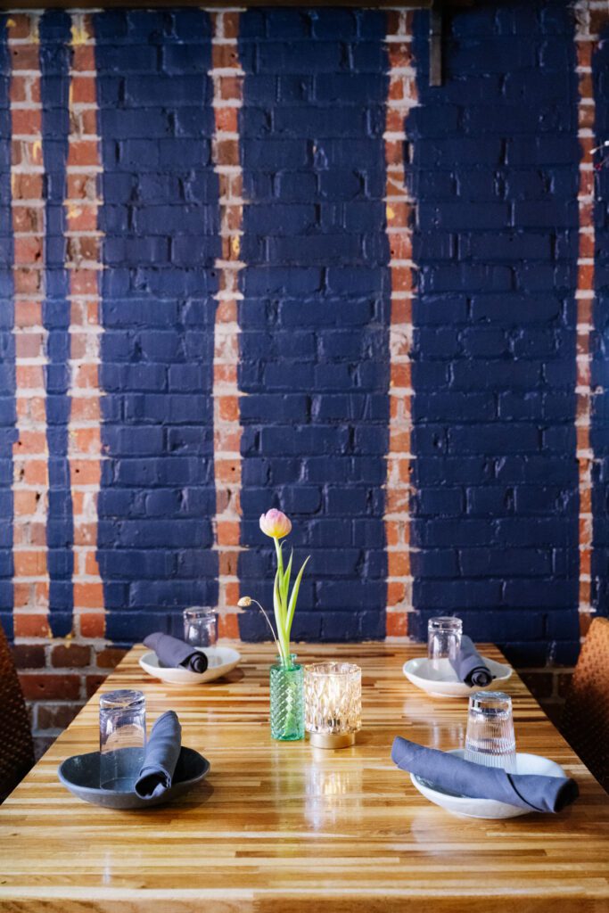 Set dining table with blue napkins and a flower centerpiece against a painted brick wall.