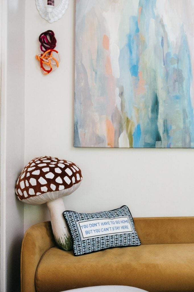 Furbish needlepoint pillow and a mushroom pillow on a mustard-colored sofa beneath abstract artwork in Courtney Hayes’ living room.