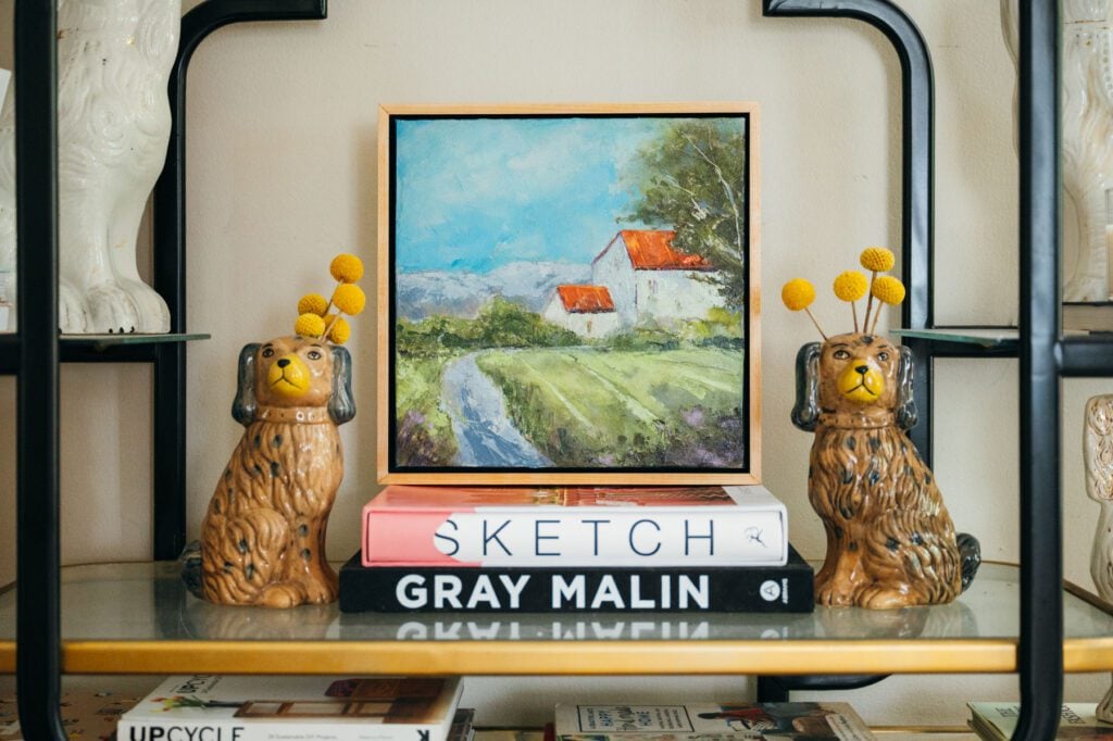 Janice Francis Art landscape painting on top of coffee table books accompanied by two ceramic English Staffordshire Dog Vases filled with yellow flowers on a bookshelf.  