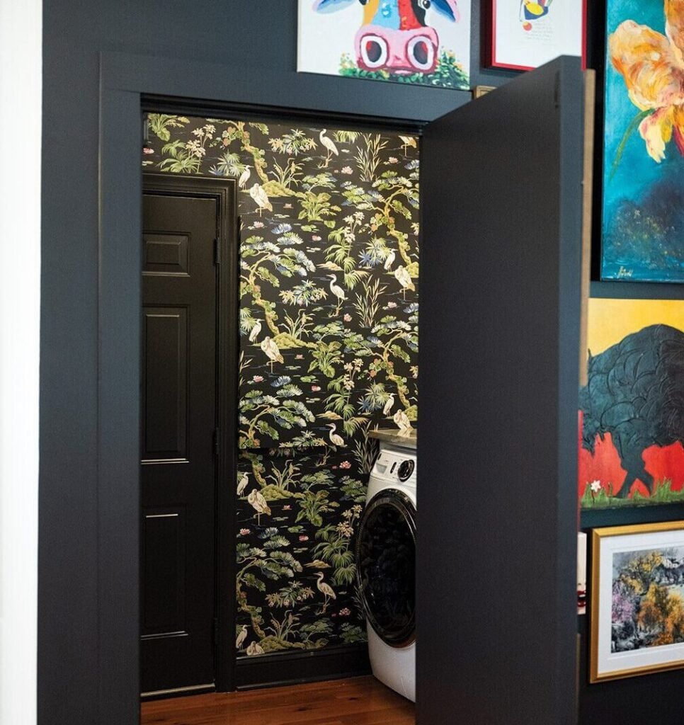 A black laundry room door (the color is Onyx by Benjamin Moore) leads to a laundry room with walls decorated whimsically bu Ronald Redding's "Bridgewater" wallpaper in black. The scene paints a dramatic look.