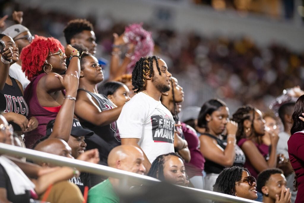 NCCU fans smile as they watch the homecoming game from the stands wearing university merchandise.