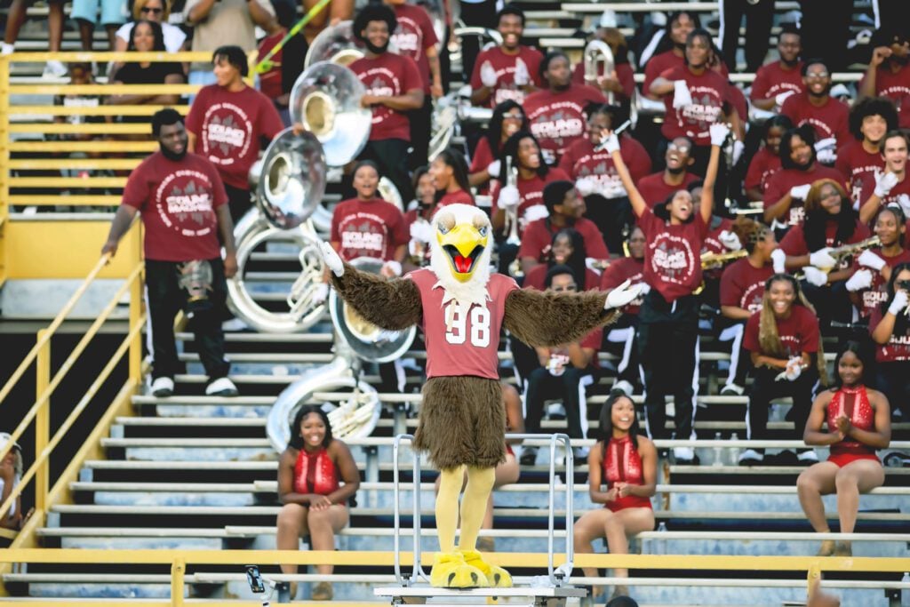NCCU mascot Eddie the Eagle wears a maroon No. 98 jersey. He stands with arms outstretched in front of a cheering marching band in stadium bleachers.